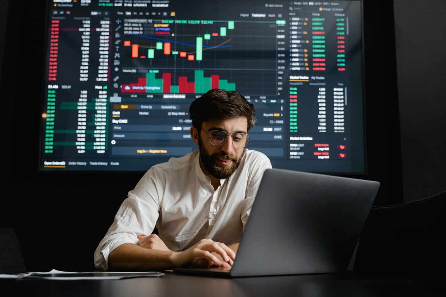 A man working on a laptop with financial charts displayed on a large screen behind him.