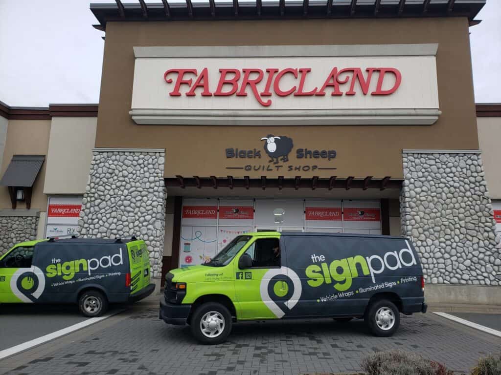 Fabricland storefront with illuminated sign and two branded vehicles parked outside.