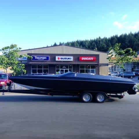 Boat on trailer parked in front of vehicle dealership with brand signs.