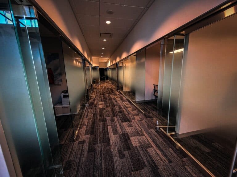 Modern glass-walled office corridor with private rooms and sleek design.