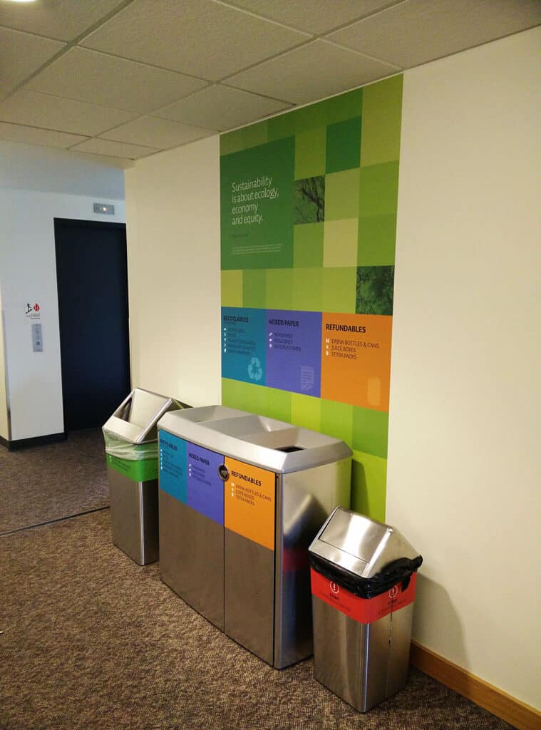 Recycling station with color-coded bins and an informational wall display in a modern office setting.