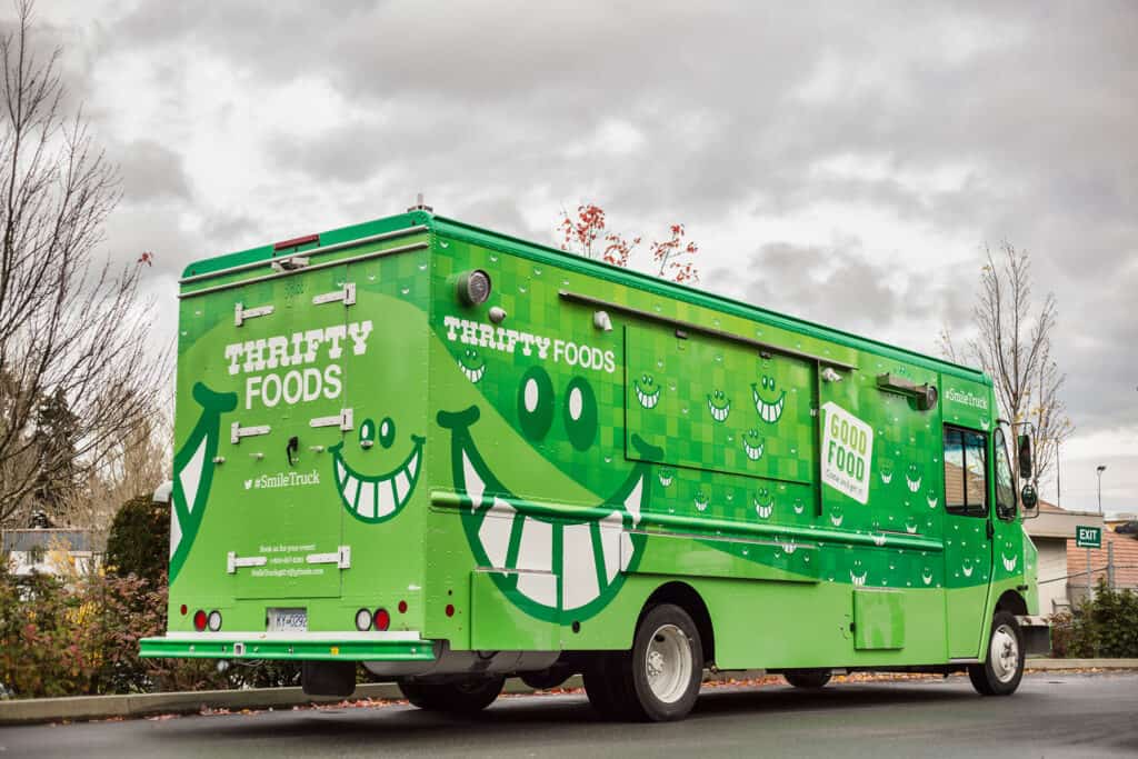 Green food truck with cheerful smiling face and "Thrifty Foods" branding, parked outdoors on a cloud.