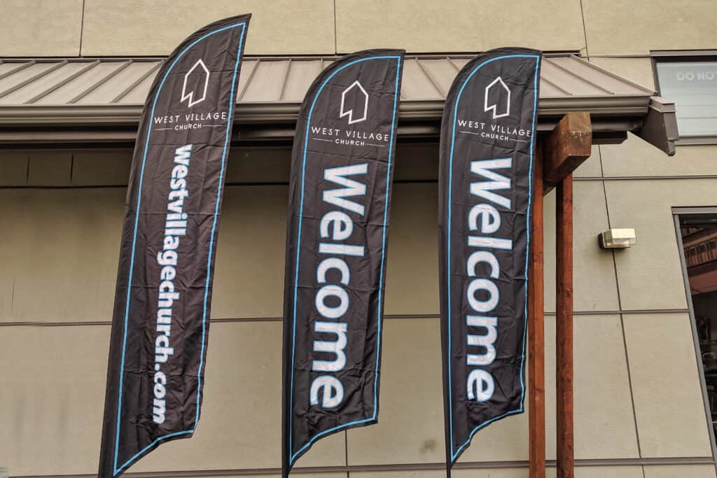 Three vertical flags with "Welcome" and church logo outside West Village Church.